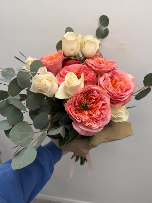A hand in a blue sleeve holds a bouquet of fresh flowers wrapped in brown paper. The arrangement features large, ruffled pink ranunculus flowers and creamy white roses, complemented by sprigs of eucal