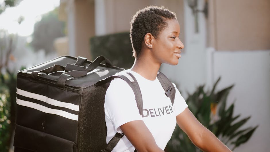 A young woman with short, textured hair and medium brown skin is seen in profile, carrying a large black insulated delivery bag with white horizontal stripes on her back. She is wearing a white T-shir